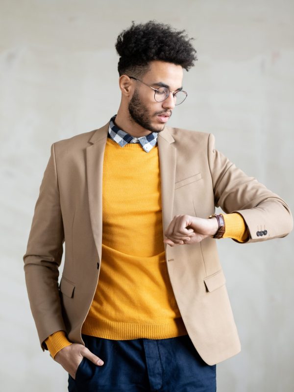 Young mixed-race serious businessman in smart casualwear standing in front of camera and looking at face of his wristwatch